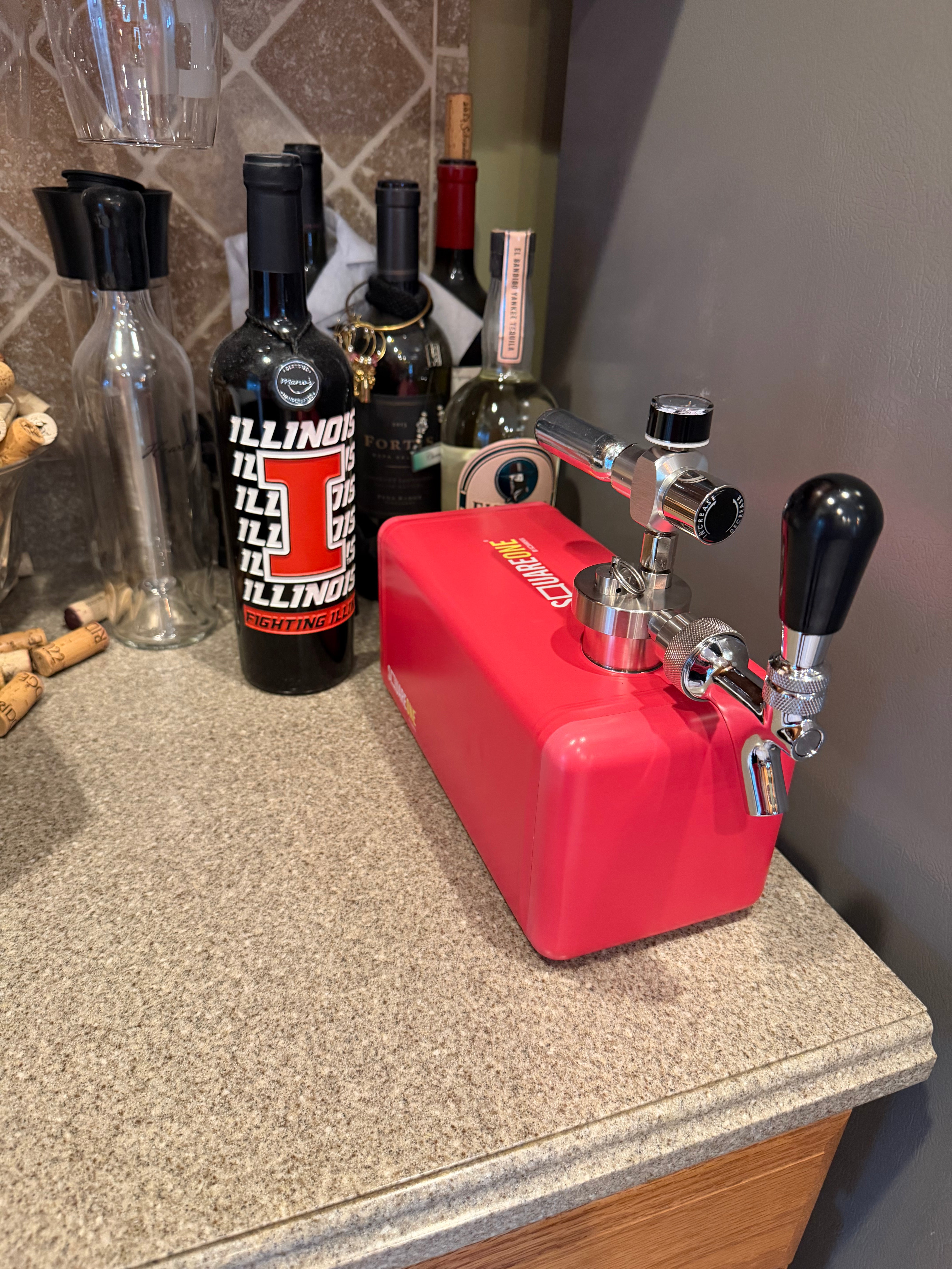 A red mini keg system with black handles, placed on a speckled granite countertop next to wine bottles.