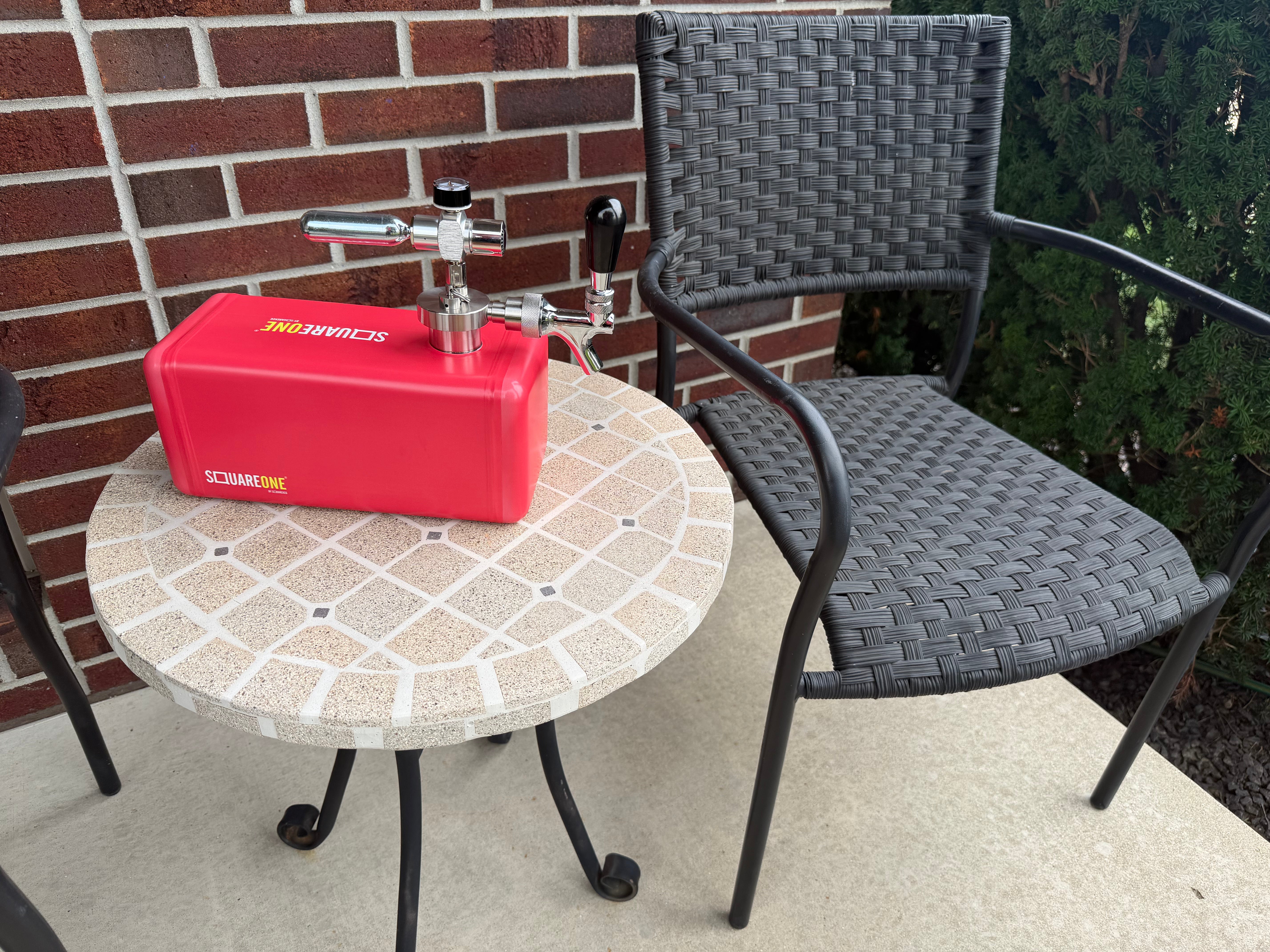 A red SquareOne keg with a stainless steel tap system sits on a round mosaic patio table next to a dark gray woven outdoor chair, set against a brick wall background.