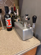 A silver SquareOne keg with chrome taps and black handles sitting on a speckled granite countertop next to several wine bottles.