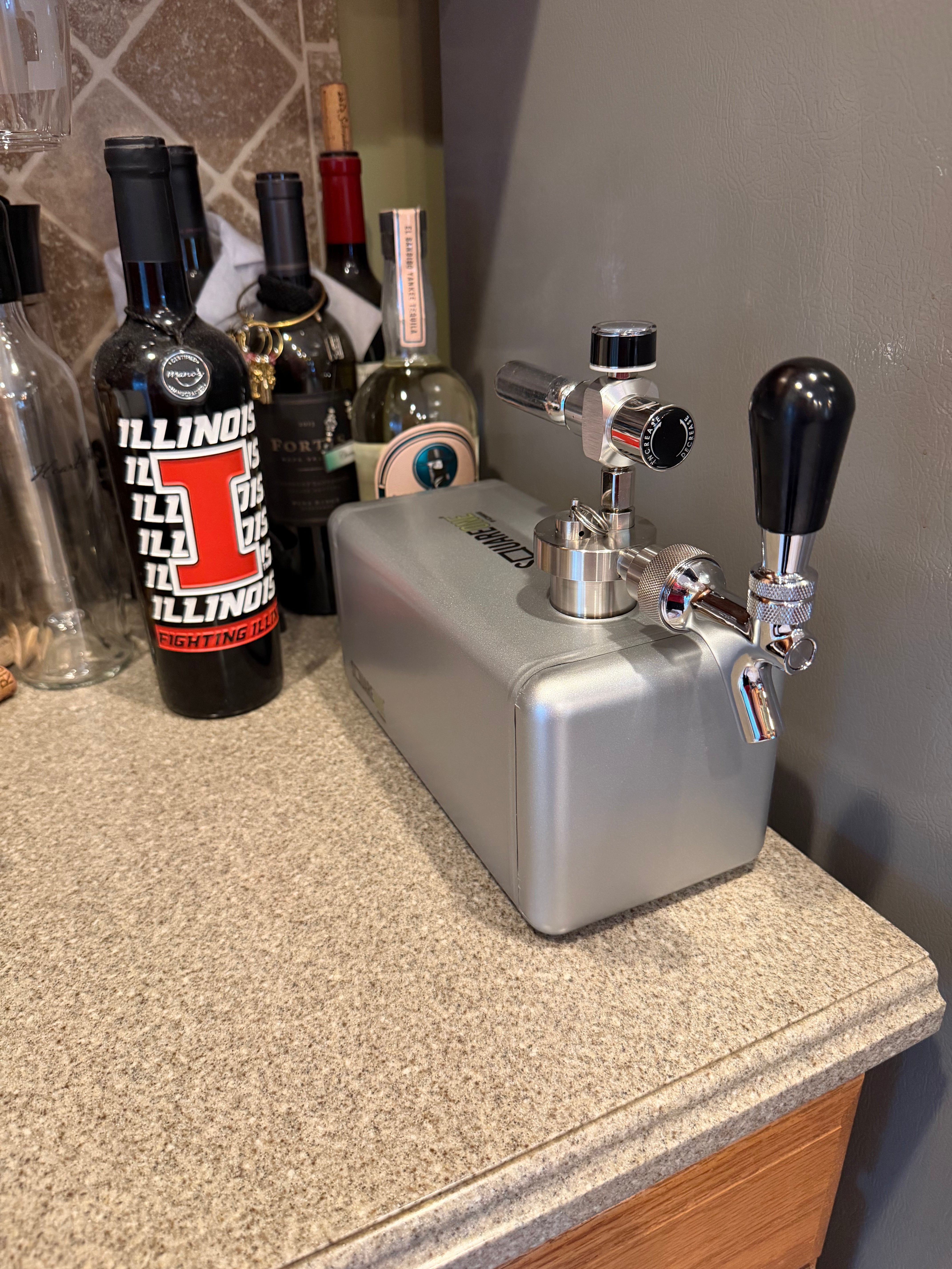 A silver SquareOne keg with chrome taps and black handles sitting on a speckled granite countertop next to several wine bottles.
